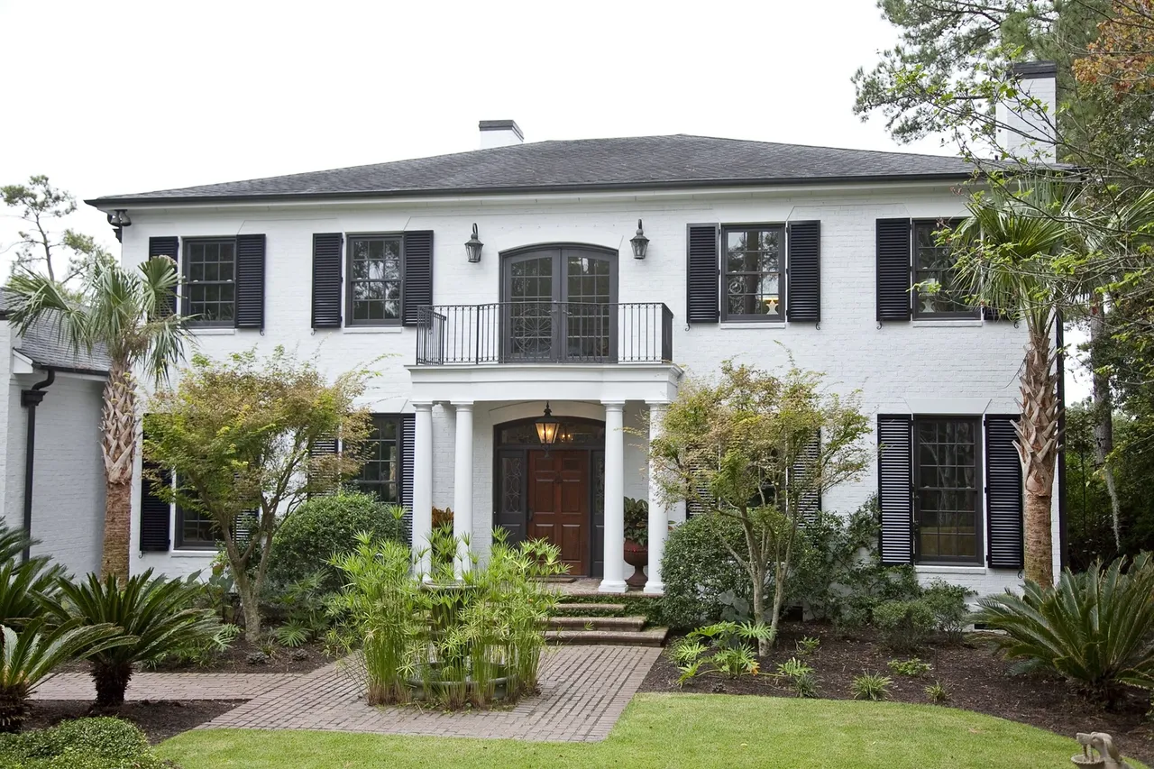 White two-story house with black shutters and lush tropical landscaping